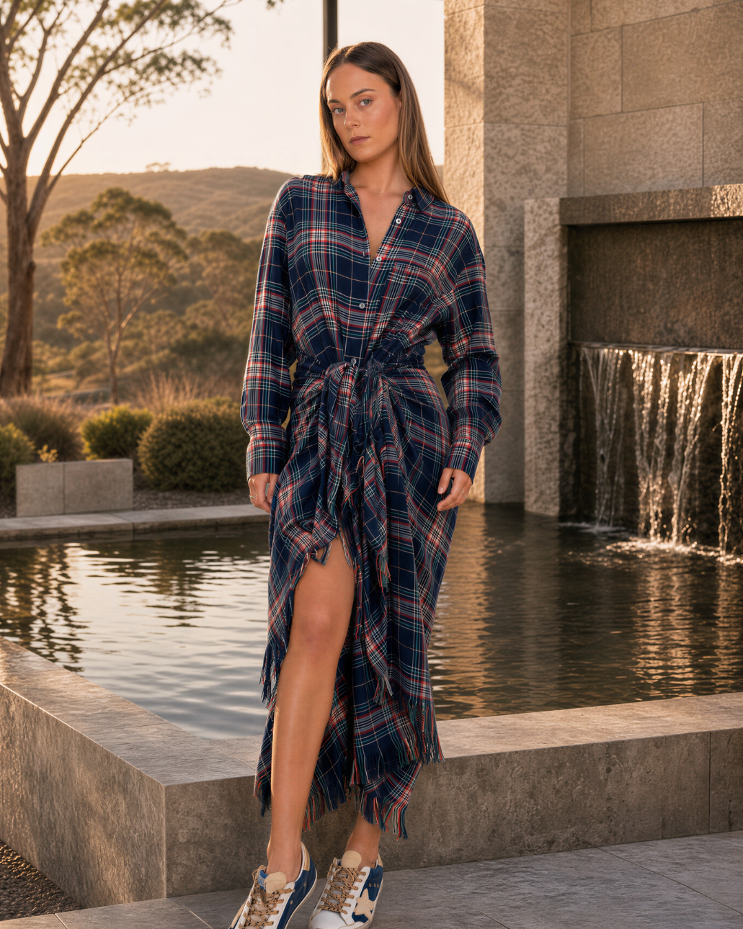 A woman models a long-sleeve plaid wrap dress with fringe details, standing in front of a stone waterfall and pool at sunset.