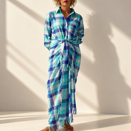 A woman modeling a blue, green, and white plaid wrap dress with a tie waist and fringed hem, standing against a warm lit studio background.