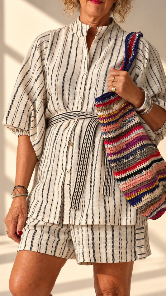 A woman wearing a cream-colored two-piece matching set with thin black vertical stripes, featuring a long-sleeve button-front top with a tie waist and matching shorts. She is holding a colorful striped knit shoulder bag.