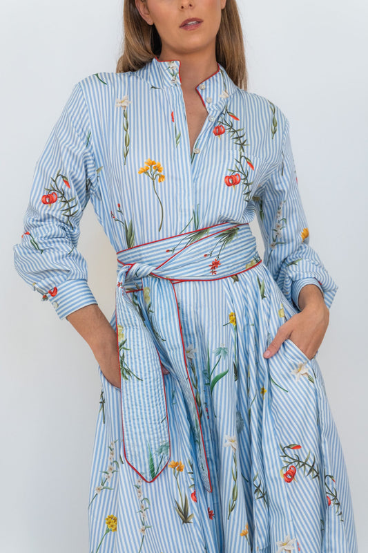 A woman wearing a light blue and white striped shirt dress with a tie waist belt and colorful floral embroidery, set against a plain white background.