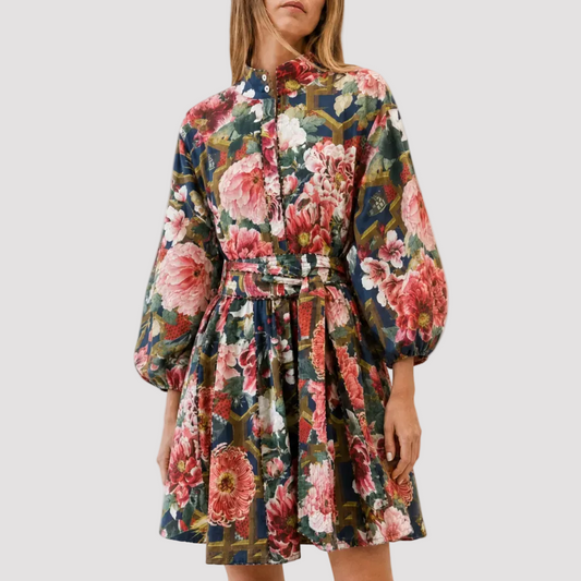 A woman modeling a floral long-sleeve mini dress with a tie waist, paired with brown leather slide sandals, against a plain white studio background.