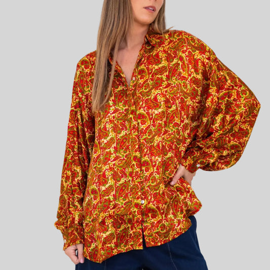 A woman wearing a loose, long-sleeve button-up blouse with an abstract red, orange, and yellow pattern, paired with dark navy pants, standing against a plain white background.