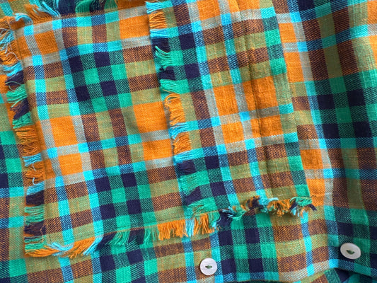 Close-up of a plaid fabric with fringe trim, featuring orange, teal, green, and dark navy checks, with white buttons visible at the top.