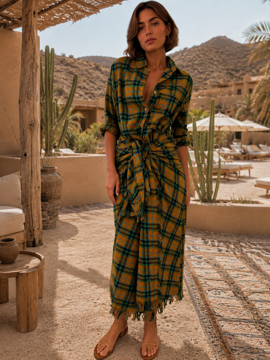 A woman models a yellow and green plaid wrap shirt dress in an outdoor desert resort setting with cacti and mountains in the background.
