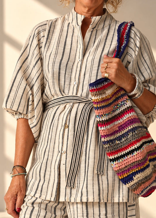 A woman wearing a cream-colored two-piece matching set with thin black vertical stripes, featuring a long-sleeve button-front top with a tie waist and matching shorts. She is holding a colorful striped knit shoulder bag.