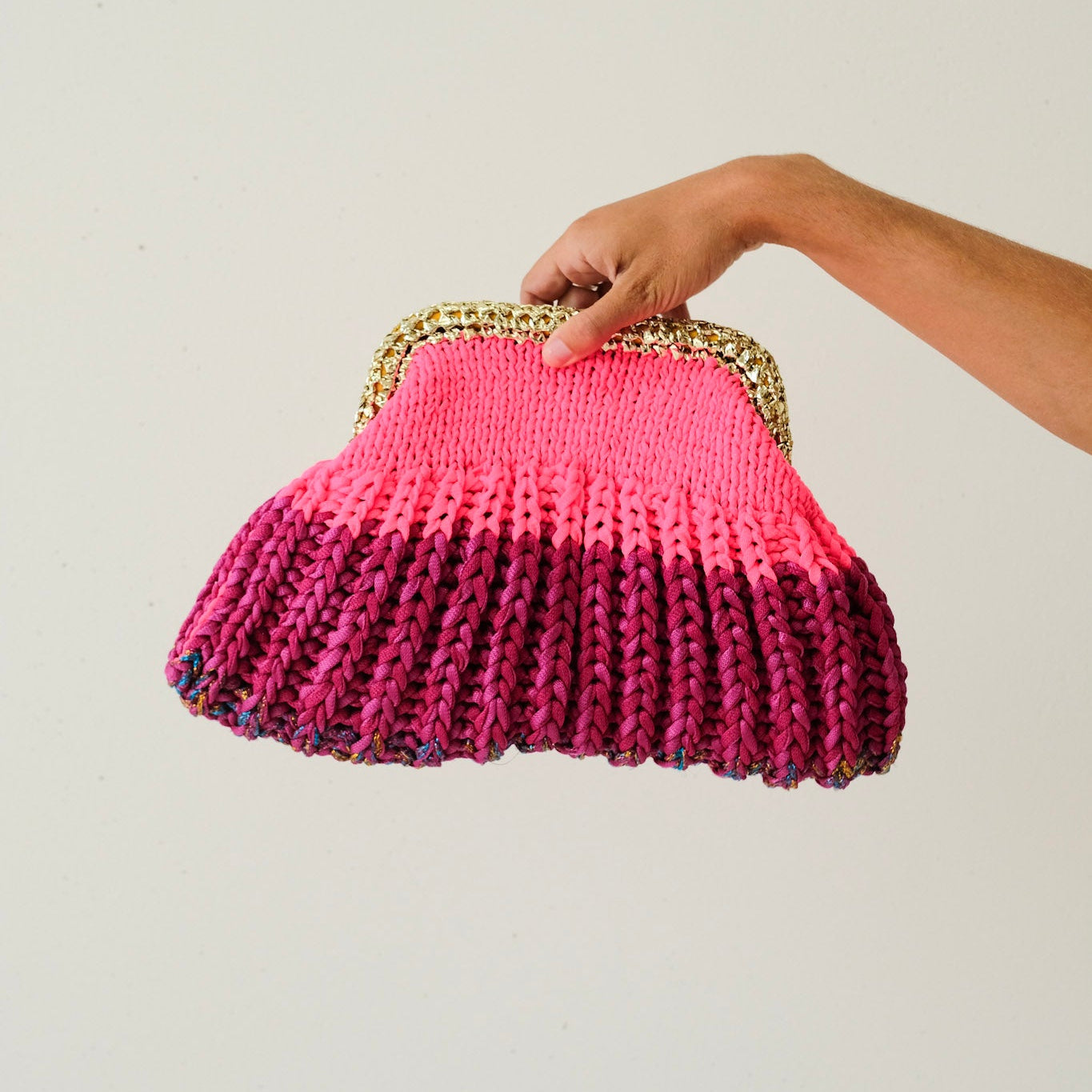 Hand holding a pink knitted clutch bag with a gold frame against a plain background