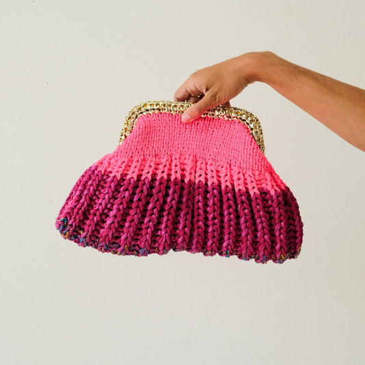 Hand holding a pink knitted clutch bag with a gold frame against a plain background
