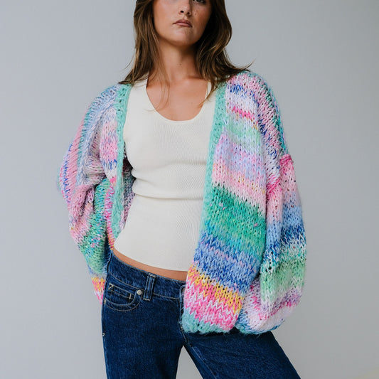 Woman wearing a colorful knitted cardigan over a white top and blue jeans on a gray background