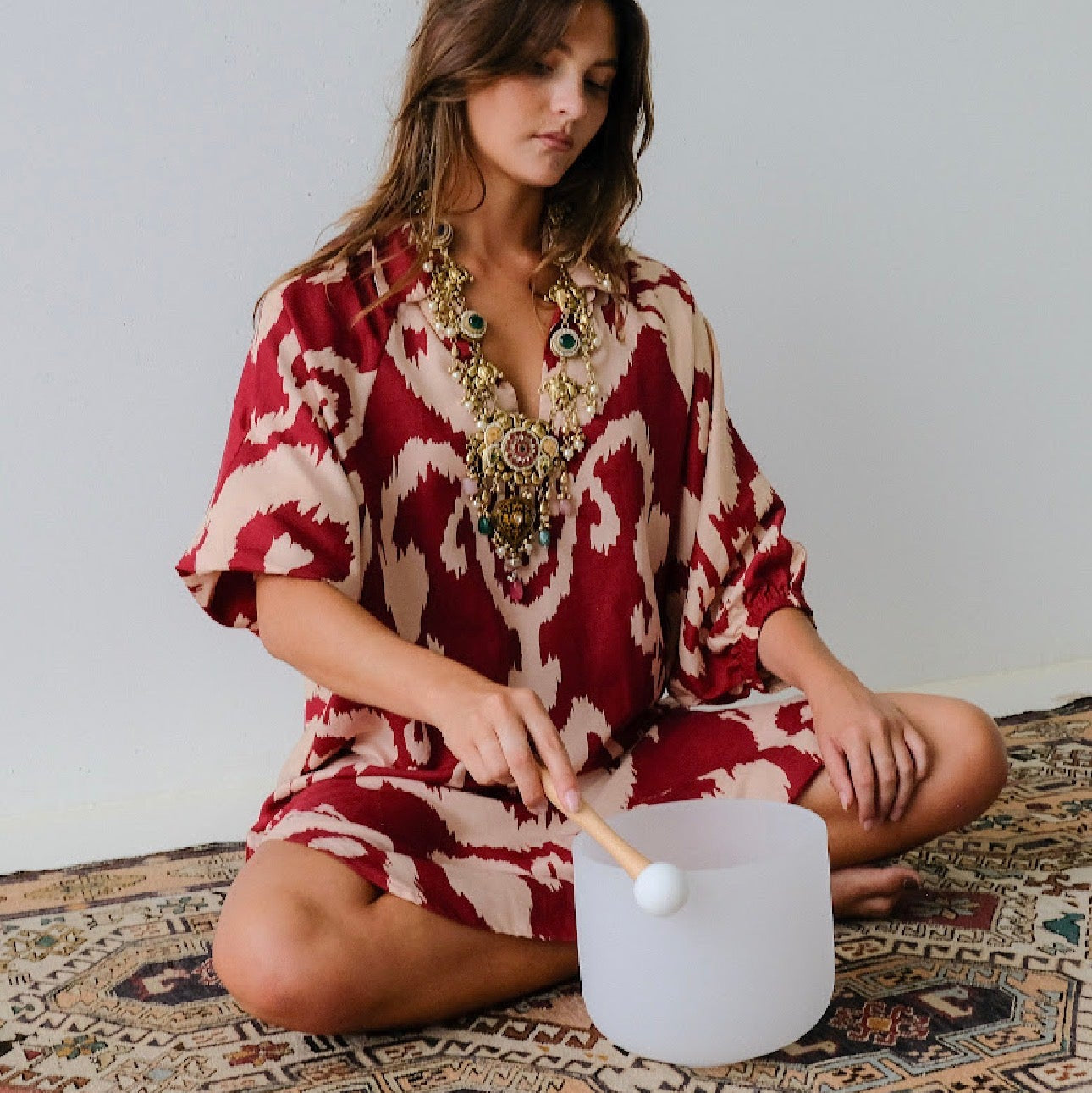 Woman in a red and white patterned dress playing a white drum on a patterned rug.