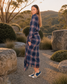 A woman wearing a navy blue plaid long-sleeve maxi dress with fringe details, standing outdoors in a rocky, landscaped area during golden hour.