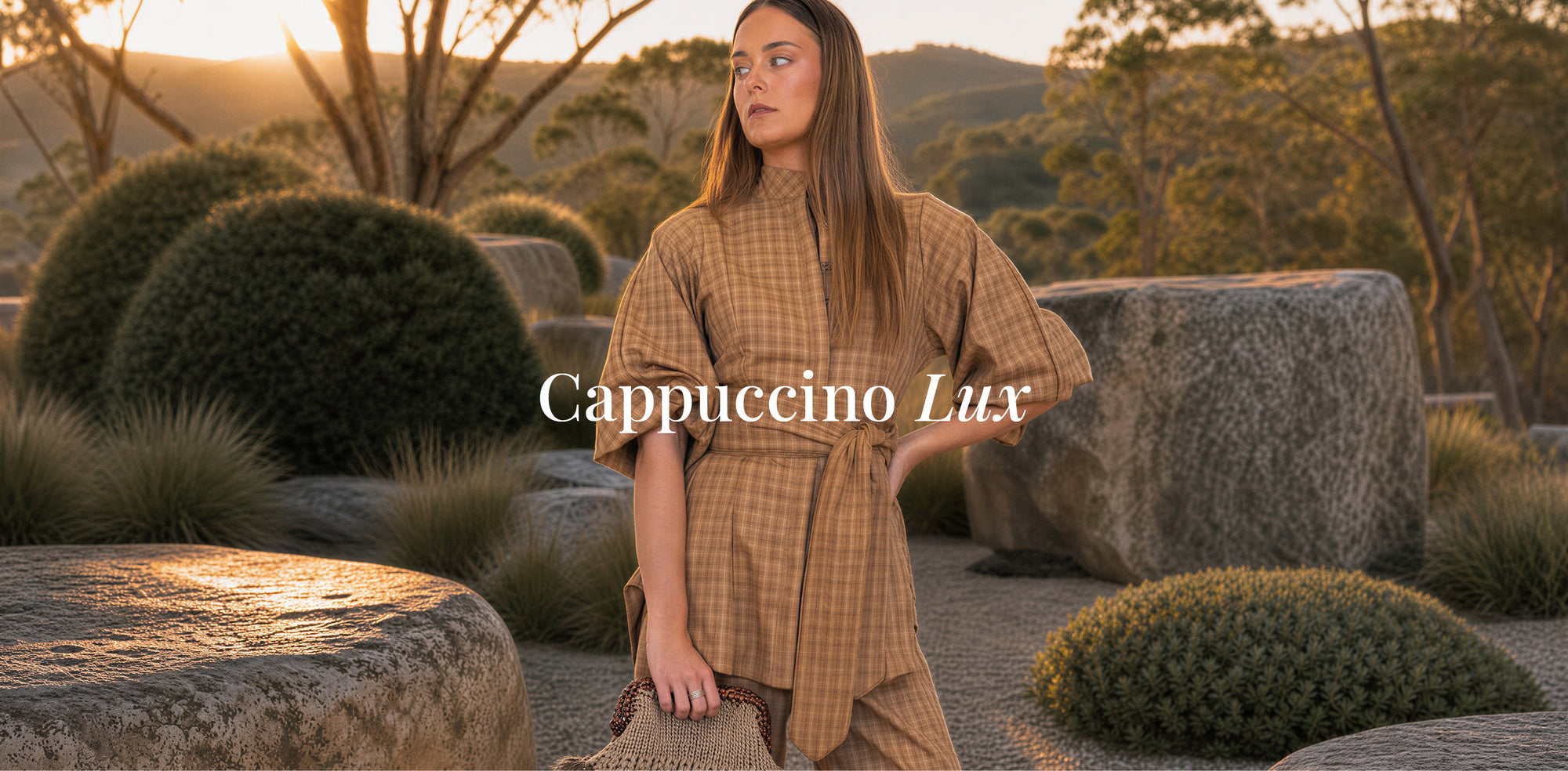 A woman stands outdoors in a rocky garden setting at sunset, wearing a loose, high-neck, long-sleeve dress in a warm tan and brown check pattern. The text 'Cappuccino Lux' is overlaid across the center.