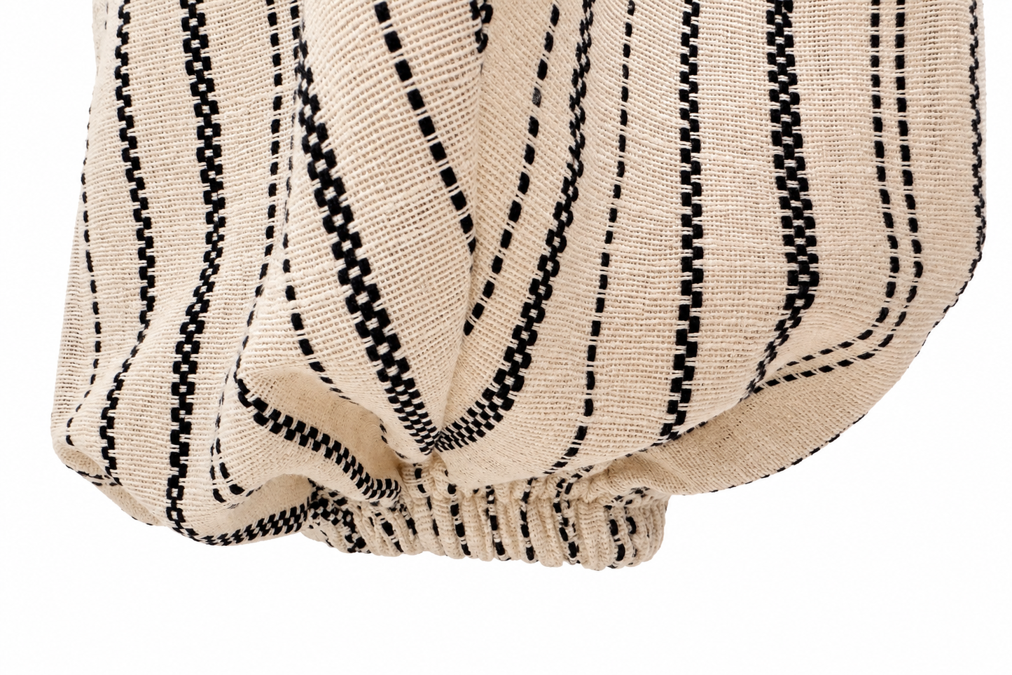 A close-up of a textured woven fabric with vertical black dotted and dashed stripes on a warm beige background, gathered at the bottom against a white backdrop.