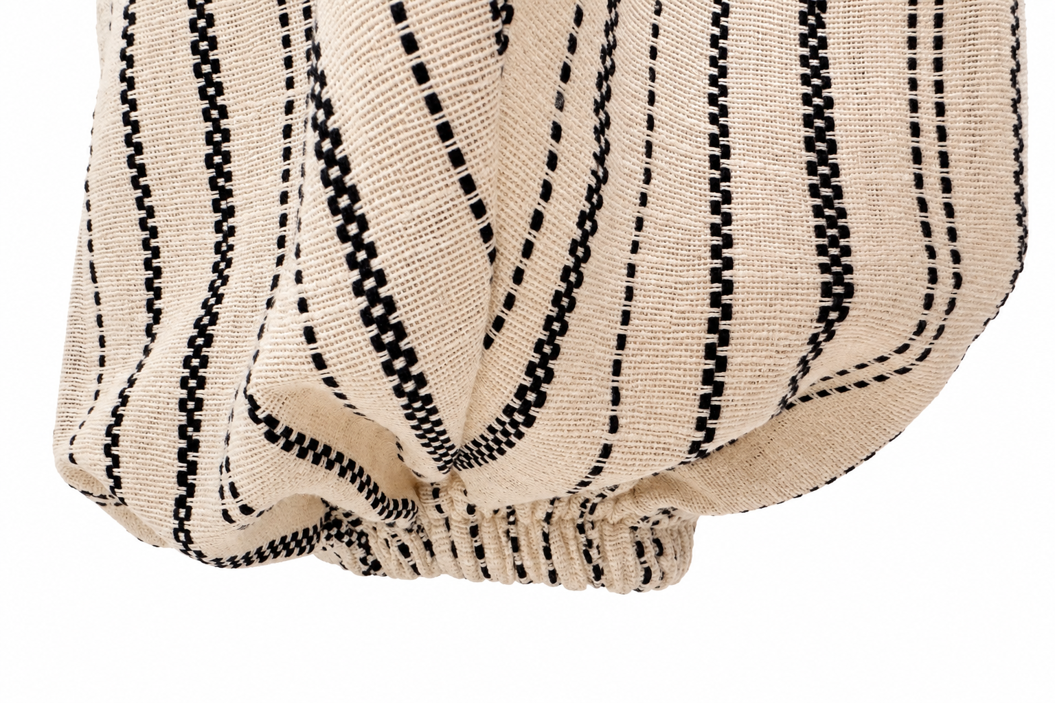 A close-up of a textured woven fabric with vertical black dotted and dashed stripes on a warm beige background, gathered at the bottom against a white backdrop.