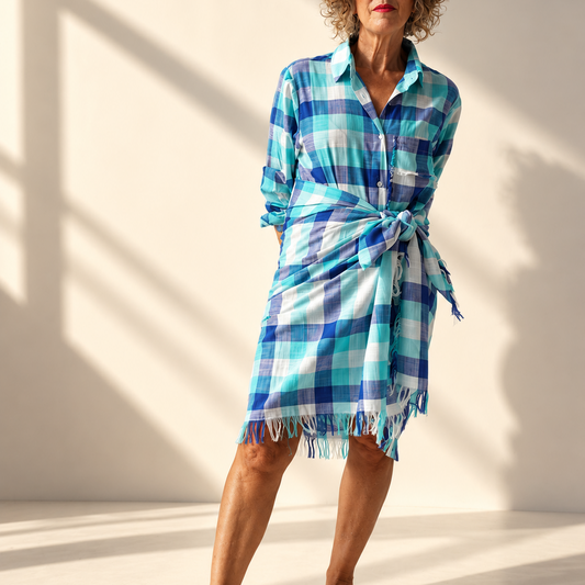A woman stands barefoot in a studio wearing a blue and white plaid wrap shirt dress with a tie waist and fringed hem, set against a warm beige wall with strong window shadows.