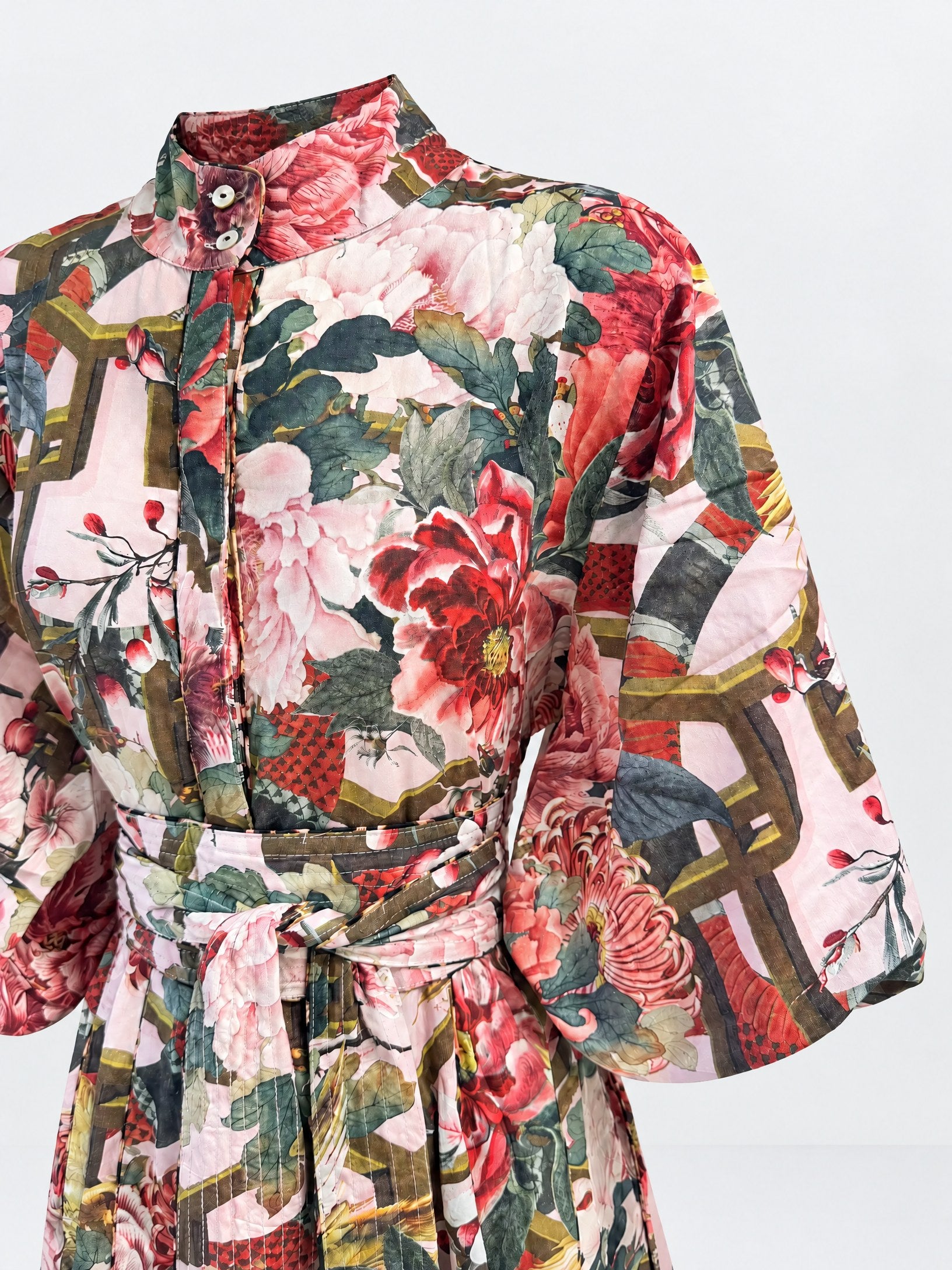 A close-up of a belted kimono-style top featuring a busy, multicolor floral and geometric print with pink, red, green, and gold tones on a light background.