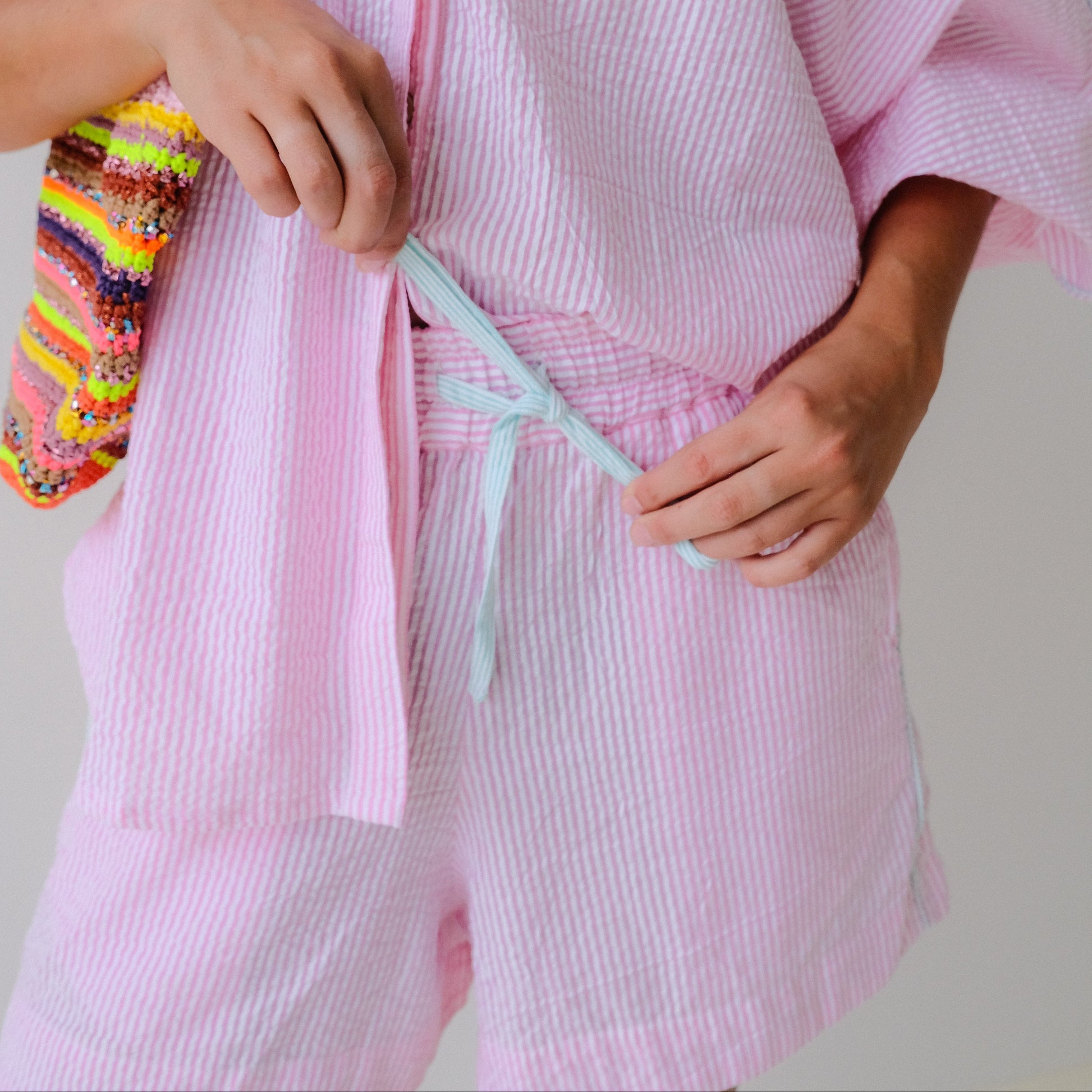 Person wearing a pink textured robe holding a colorful bag and a light blue item.