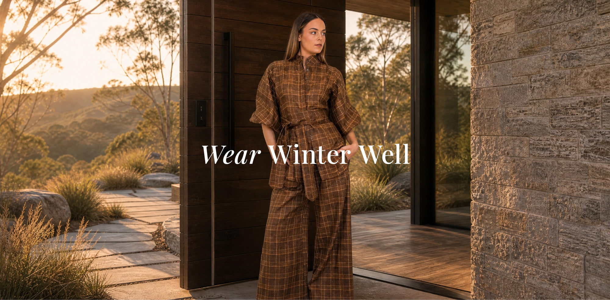 A woman wearing a brown and beige plaid matching set stands in a doorway of a modern house, with a warm outdoor landscape in the background. The text 'Wear Winter Well' is overlaid on the center.