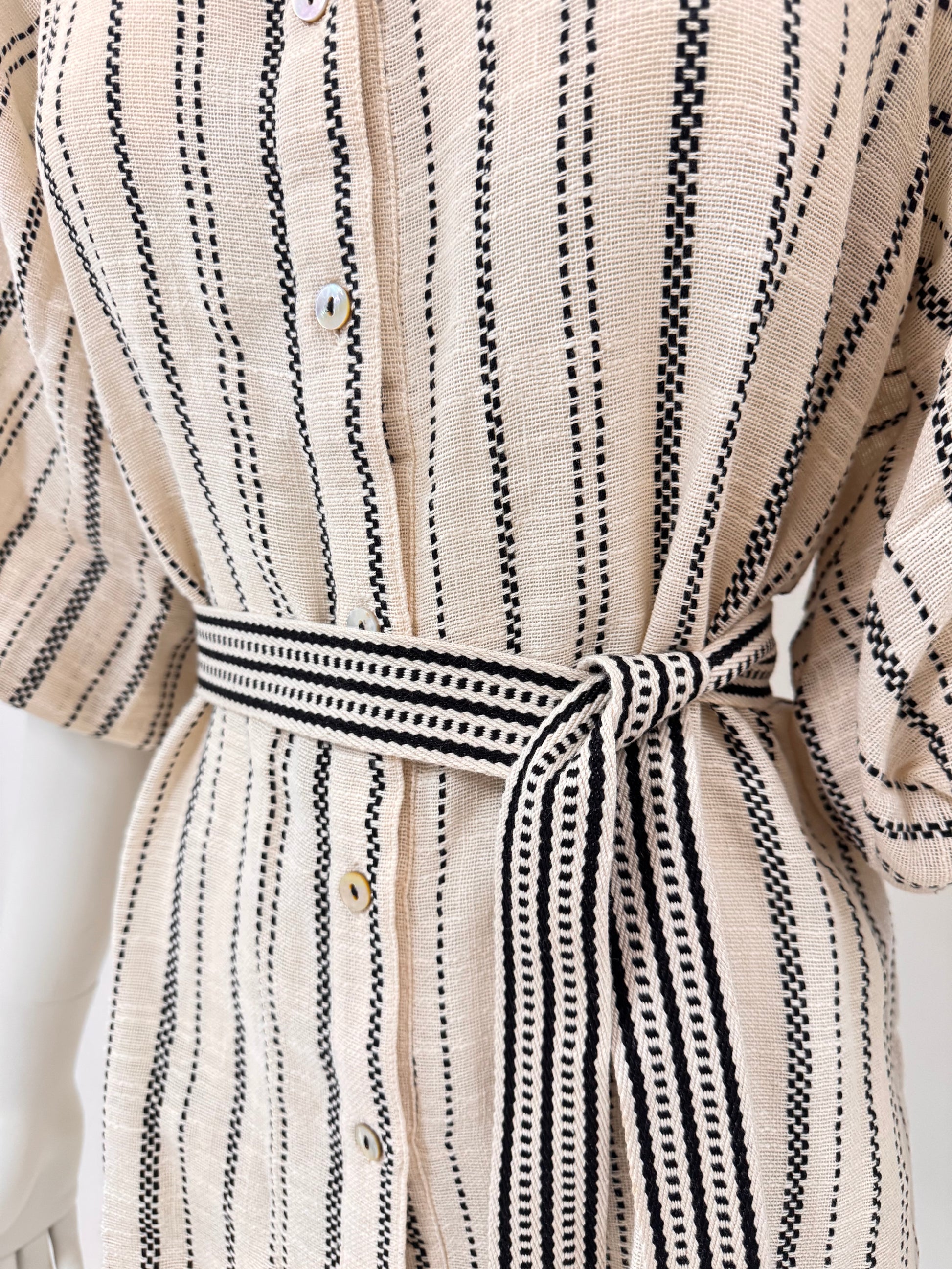 Close-up of a beige woven shirt dress with black vertical dotted stripe patterns and a matching self-tie belt at the waist.