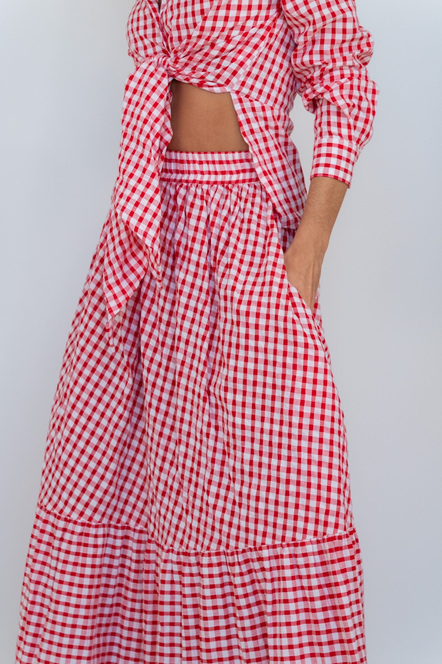 A red and white gingham two-piece set featuring a long-sleeve crop top with a side tie and a matching tiered maxi skirt with side pockets, shown on a model against a light gray background.