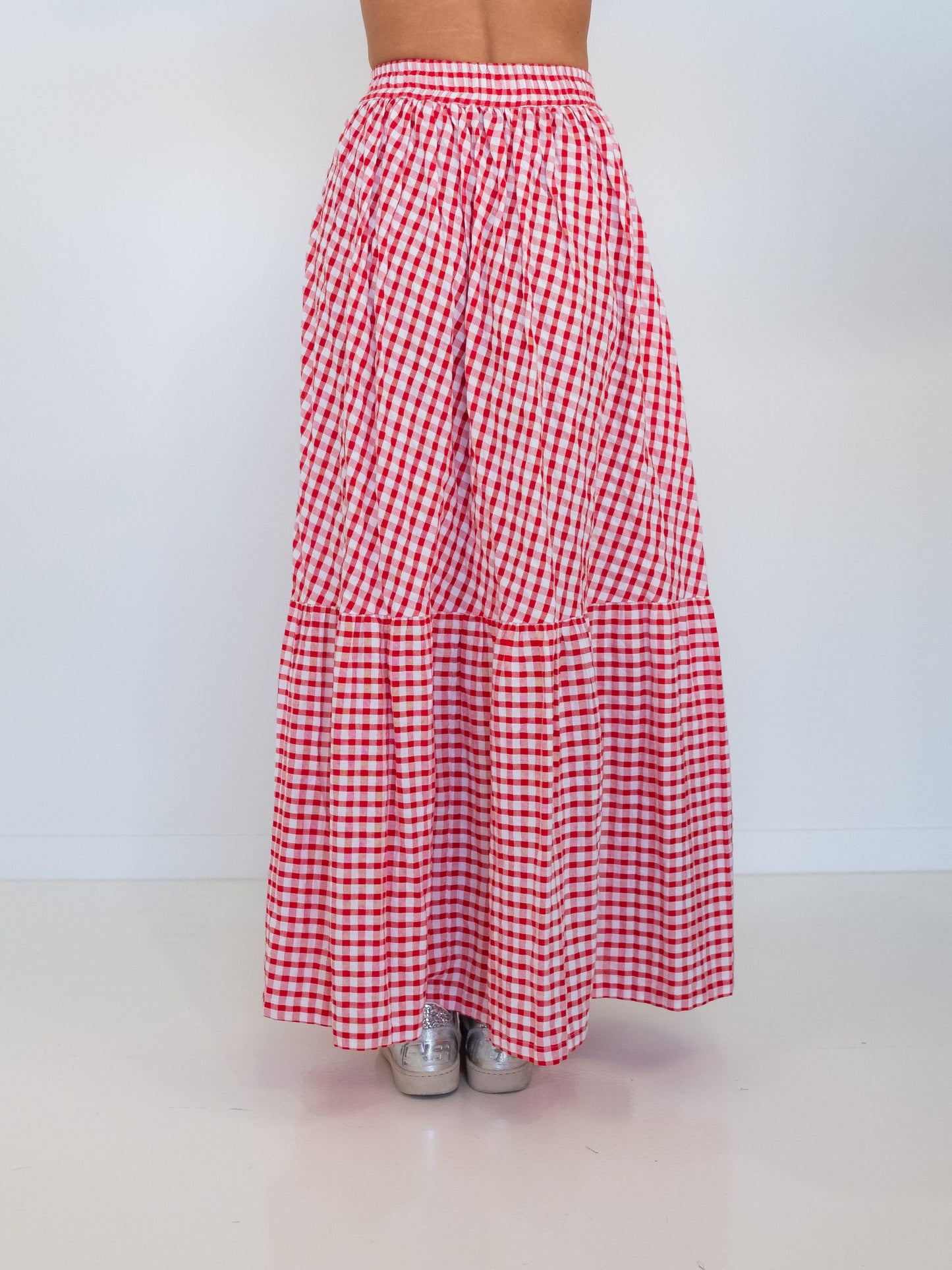 A red and white gingham tiered maxi skirt shown from the back on a model, paired with silver sneakers, against a plain white studio background.