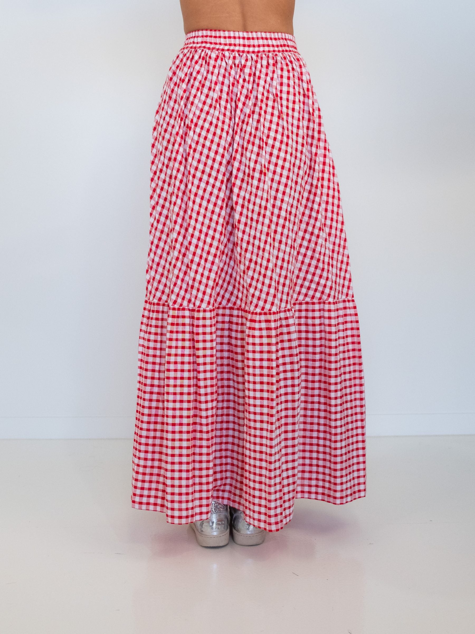 A red and white gingham tiered maxi skirt shown from the back on a model, paired with silver sneakers, against a plain white studio background.