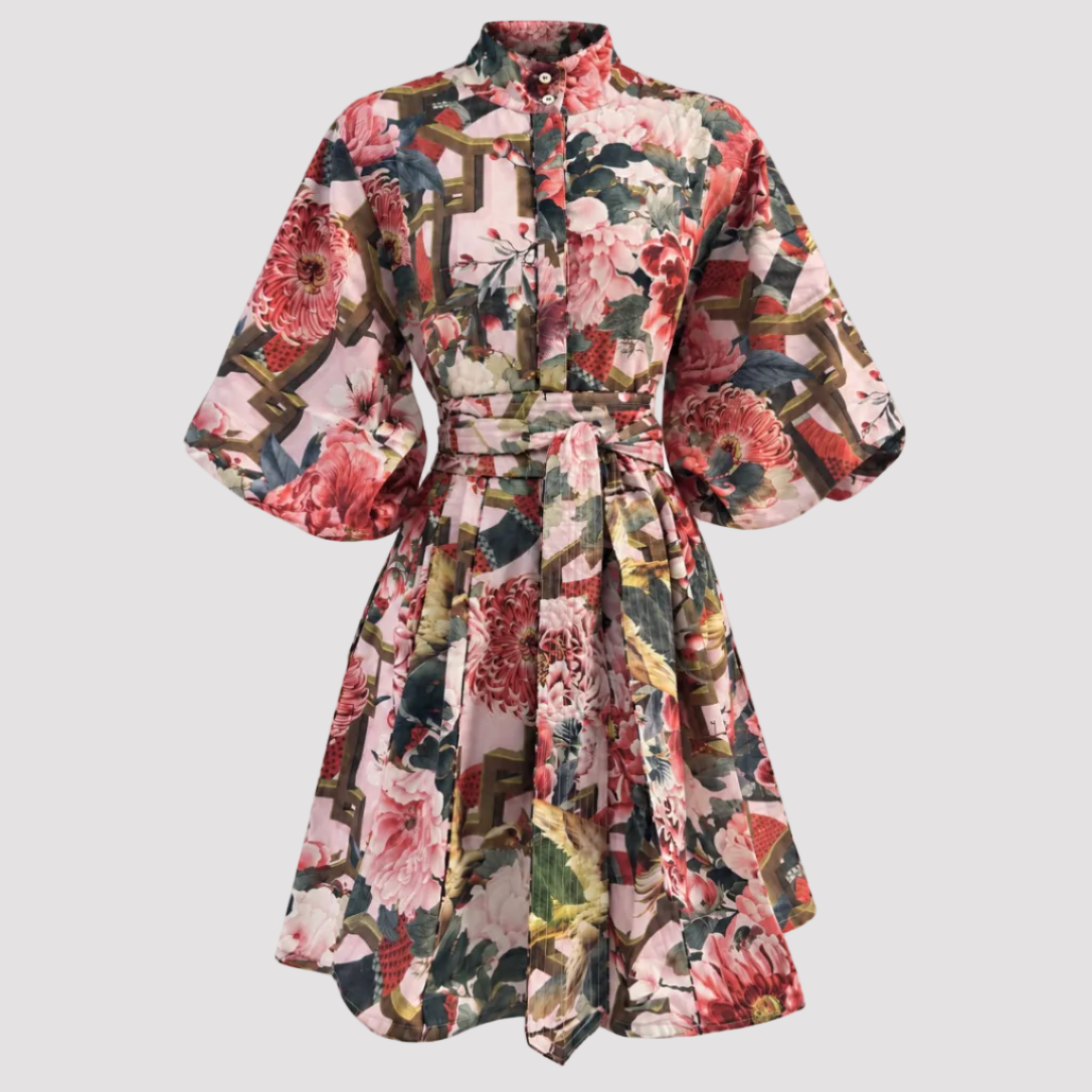 A short-sleeved, button-front dress with a self-tie waist belt, featuring a dense floral and architectural print in pink, red, green, and beige tones, shown against a plain white background.