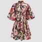 A short-sleeved, button-front dress with a self-tie waist belt, featuring a dense floral and architectural print in pink, red, green, and beige tones, shown against a plain white background.