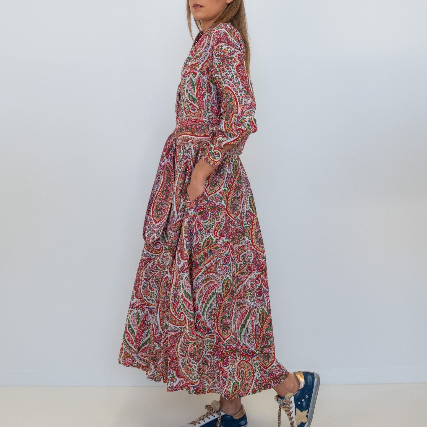 A woman modeling a long-sleeved maxi dress with a red, green, and beige paisley pattern, paired with blue sneakers, against a plain white studio background.
