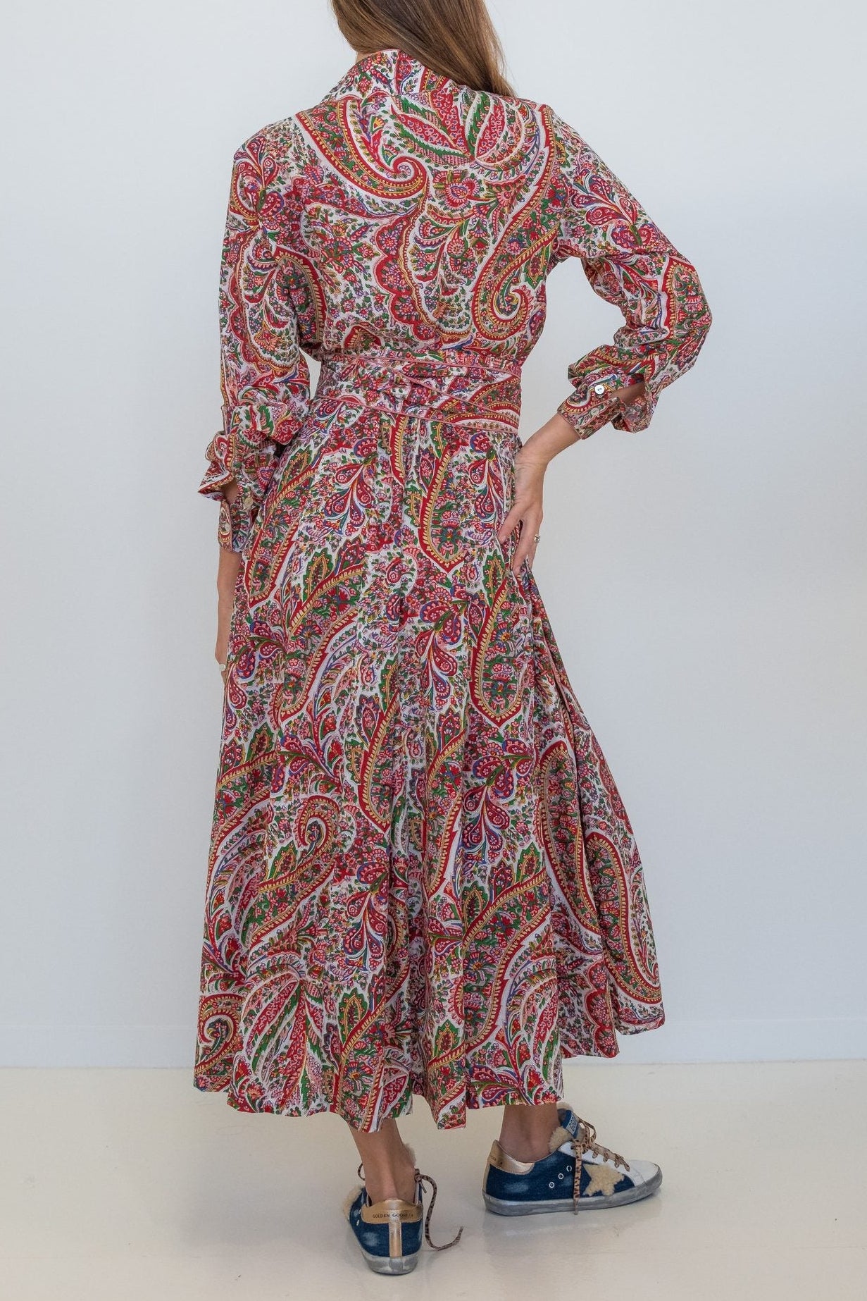 A woman viewed from behind wearing a long-sleeved, collared maxi dress with a red, pink, and green paisley pattern, paired with blue sneakers, against a plain white studio background.