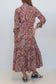 A woman viewed from behind wearing a long-sleeved, collared maxi dress with a red, pink, and green paisley pattern, paired with blue sneakers, against a plain white studio background.