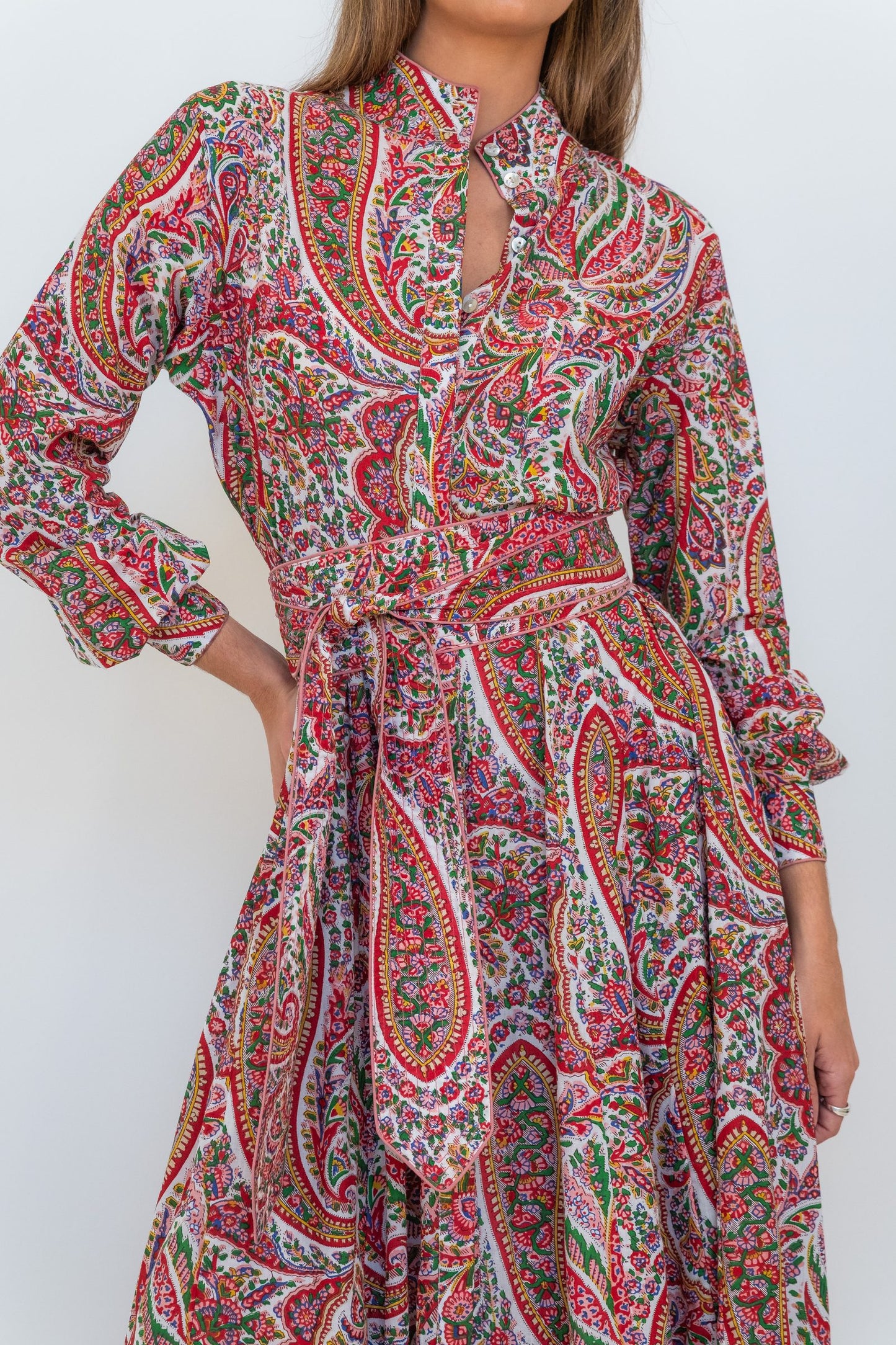 A woman wearing a long-sleeve, high-neck dress with a vibrant red, green, and orange paisley pattern on a white base, featuring a keyhole cutout at the collar and a matching waist tie.