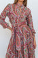 A woman wearing a long-sleeve, high-neck dress with a vibrant red, green, and orange paisley pattern on a white base, featuring a keyhole cutout at the collar and a matching waist tie.