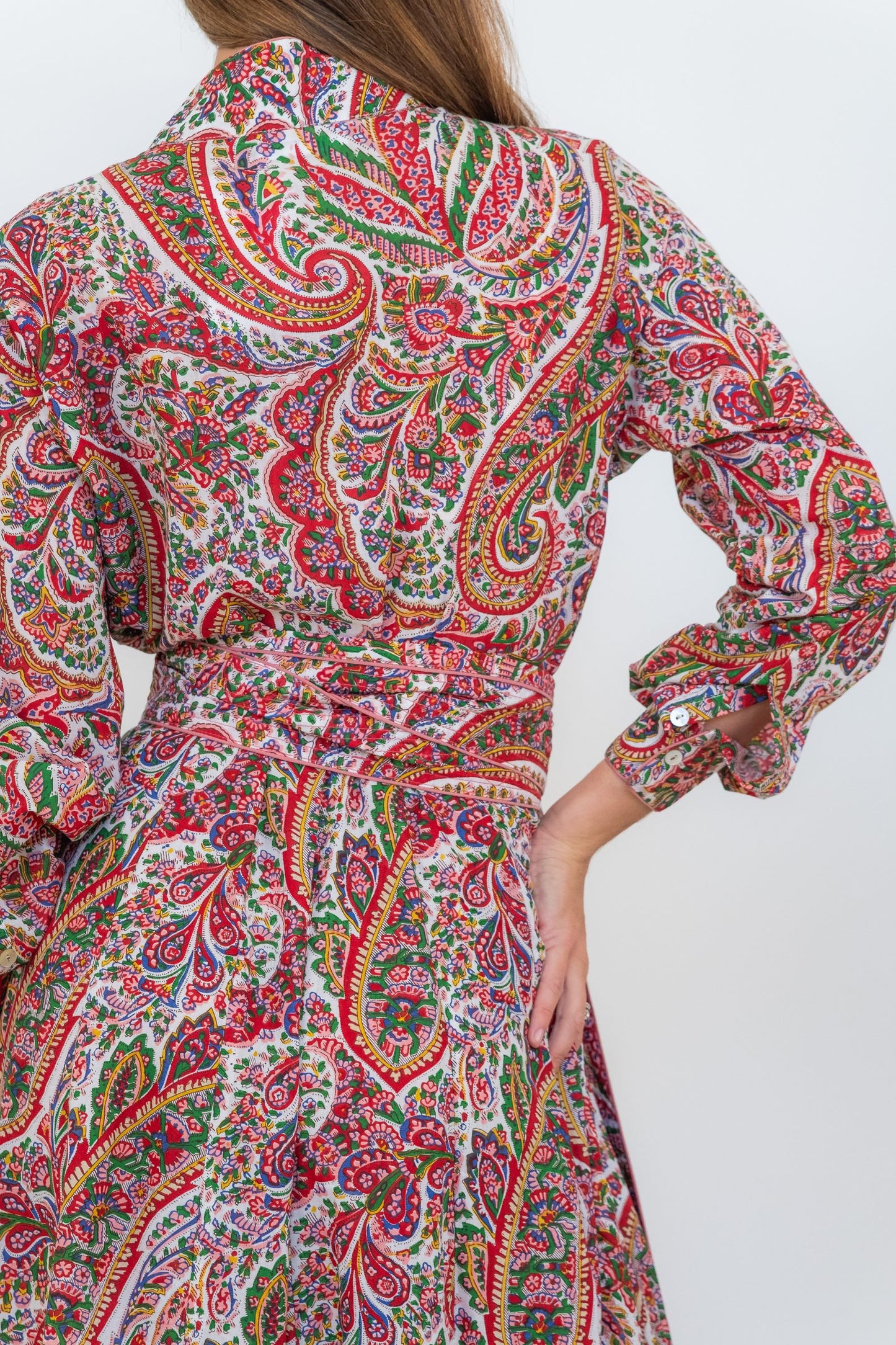 A close-up of a model wearing a long-sleeved collared dress with a vibrant red, green, and blue paisley pattern on a cream background.