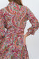A close-up of a model wearing a long-sleeved collared dress with a vibrant red, green, and blue paisley pattern on a cream background.