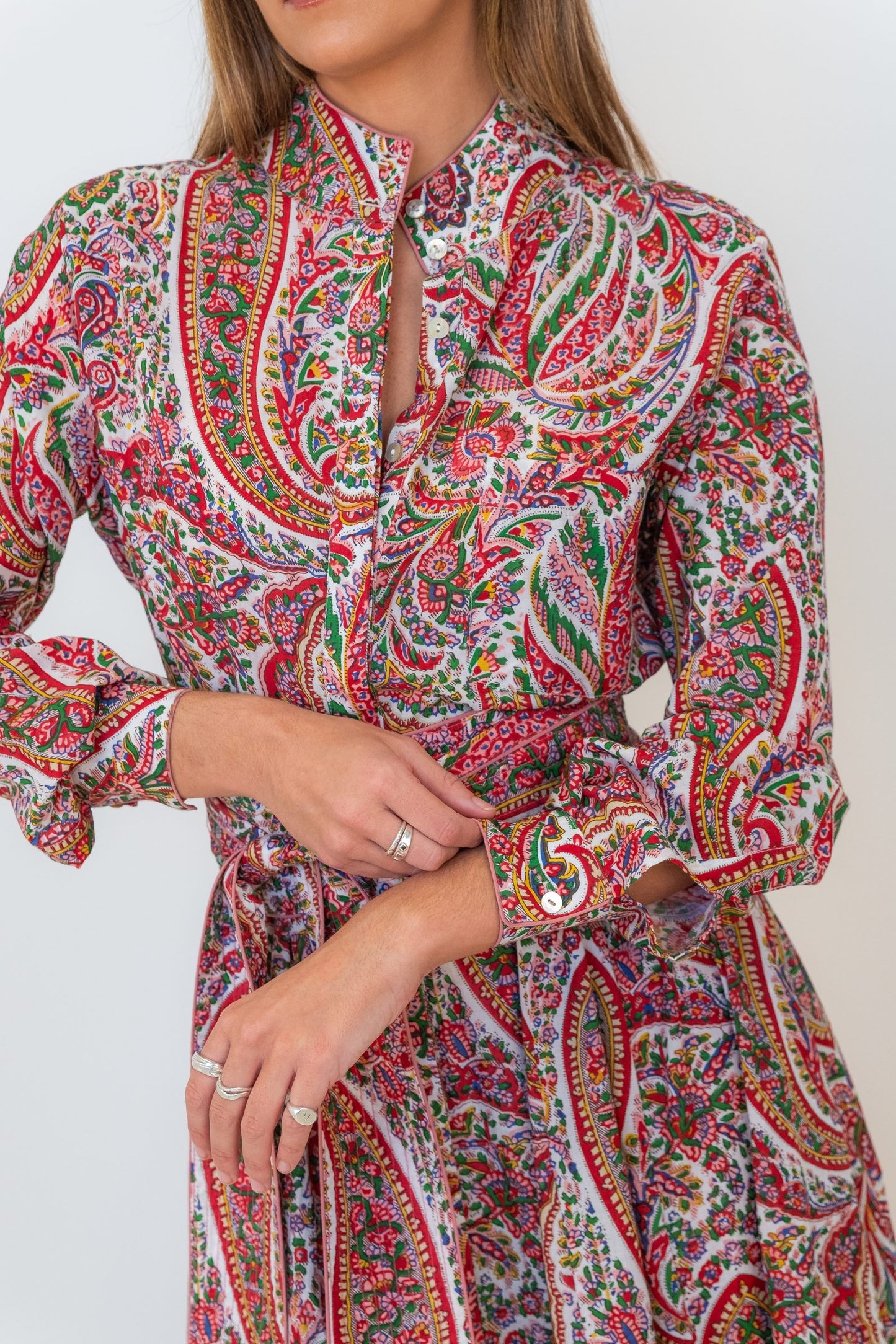 A close-up of a woman wearing a long-sleeve collared dress with a dense paisley print in red, green, blue, and yellow on a light background.