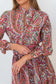 A close-up of a woman wearing a long-sleeve collared dress with a dense paisley print in red, green, blue, and yellow on a light background.