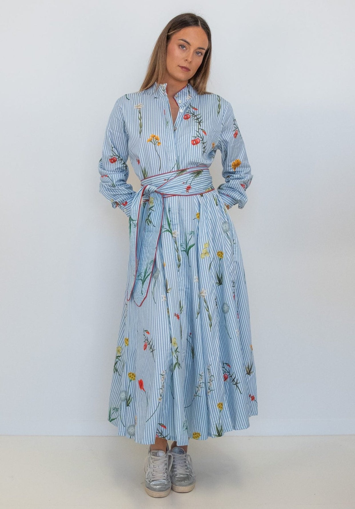 A woman modeling a light blue long-sleeve maxi shirt dress with a colorful floral and bird print, featuring a matching tie belt, set against a plain white studio background.