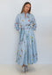 A woman modeling a light blue long-sleeve maxi shirt dress with a colorful floral and bird print, featuring a matching tie belt, set against a plain white studio background.