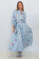 A woman modeling a light blue long-sleeve maxi shirt dress with a vertical stripe pattern and a colorful floral print, styled with gray sneakers against a plain white studio background.