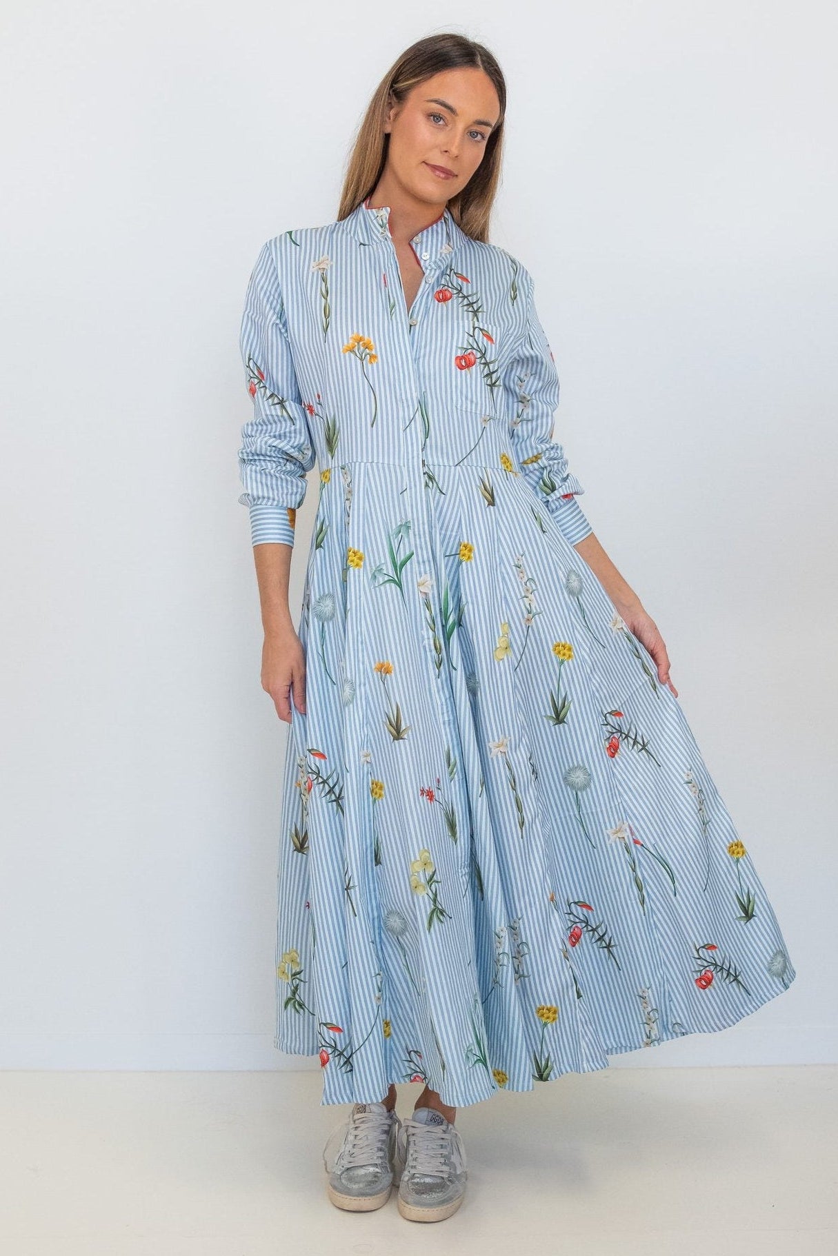 A woman modeling a light blue long-sleeve maxi dress with a vertical stripe pattern and a colorful floral print, paired with gray sneakers, against a plain white studio background.