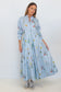 A woman modeling a light blue long-sleeve maxi dress with a vertical stripe pattern and a colorful floral print, paired with gray sneakers, against a plain white studio background.