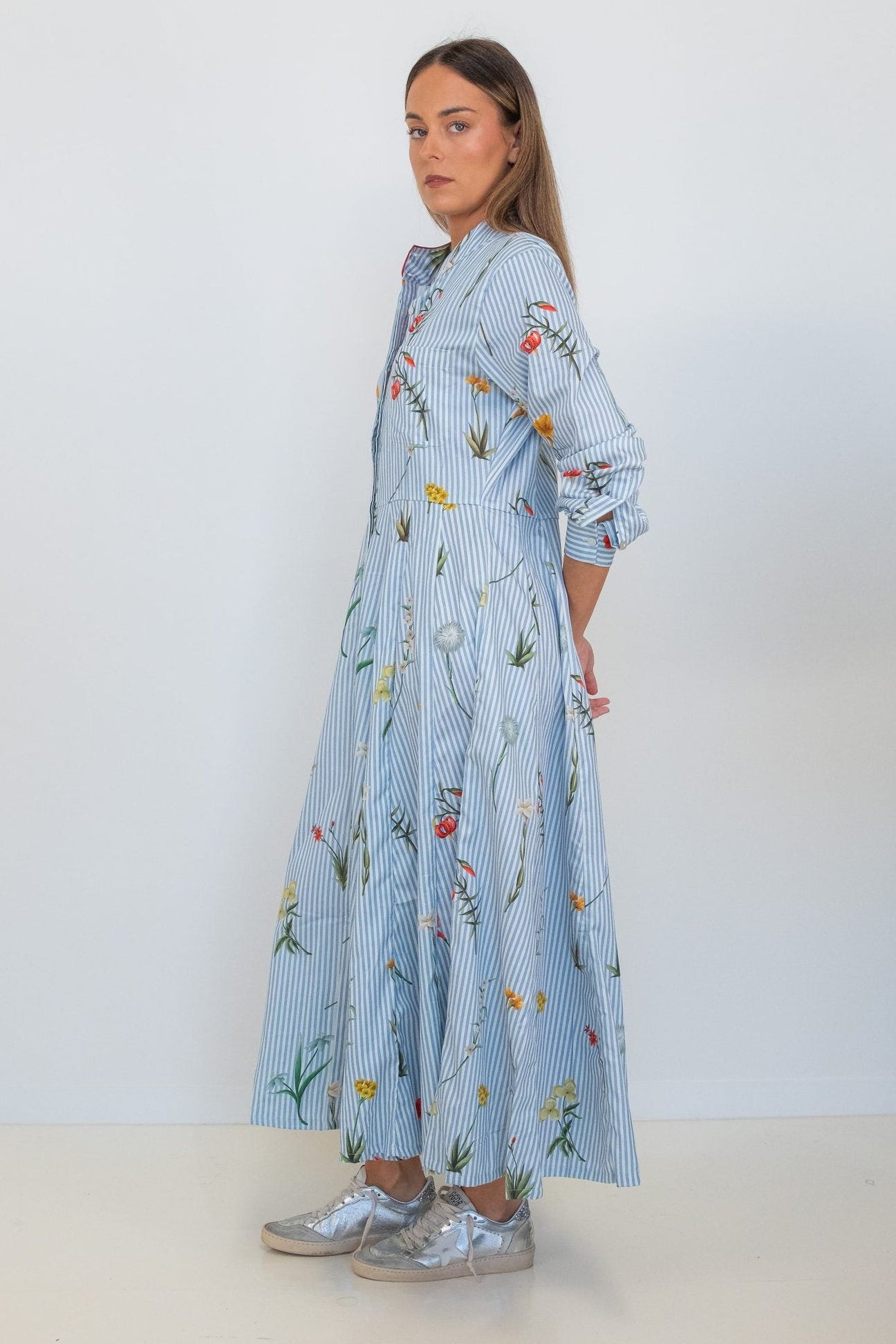 A woman modeling a light blue striped shirt dress with a colorful wildflower print, paired with silver sneakers, standing against a plain white studio background.