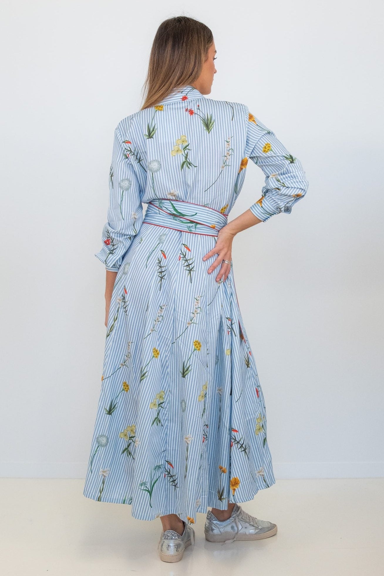 A woman is shown from the back wearing a light blue and white vertically striped long-sleeve midi dress with a floral print and a matching tie belt, paired with silver sneakers against a plain white studio background.