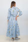 A woman is shown from the back wearing a light blue and white vertically striped long-sleeve midi dress with a floral print and a matching tie belt, paired with silver sneakers against a plain white studio background.