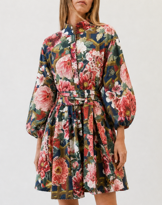A woman modeling a floral long-sleeve mini dress with a tie waist, paired with brown leather slide sandals, against a plain white studio background.