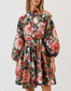 A woman modeling a floral long-sleeve mini dress with a tie waist, paired with brown leather slide sandals, against a plain white studio background.