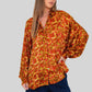 A woman wearing a loose, long-sleeve button-up blouse with an abstract red, orange, and yellow pattern, paired with dark navy pants, standing against a plain white background.