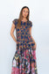 A woman modeling a tiered maxi dress with a navy blue base featuring a gold chain-link pattern on the bodice and a colorful floral print on the skirt, set against a plain white studio background.