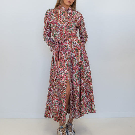 A woman modeling a long-sleeved, high-neck maxi dress with a red, pink, and blue paisley pattern, standing against a plain white studio background.