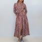 A woman modeling a long-sleeved, high-neck maxi dress with a red, pink, and blue paisley pattern, standing against a plain white studio background.